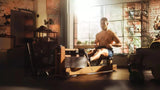 Man rowing on premium wooden rowing machine in a sunlit home gym with industrial-style decor, promoting Luna.Fitness's message 'Empower Your Fitness Journey with Luna.Fitness' and showcasing high-quality workout equipment designed for achieving fitness goals at home. Call-to-action: Explore All Gear